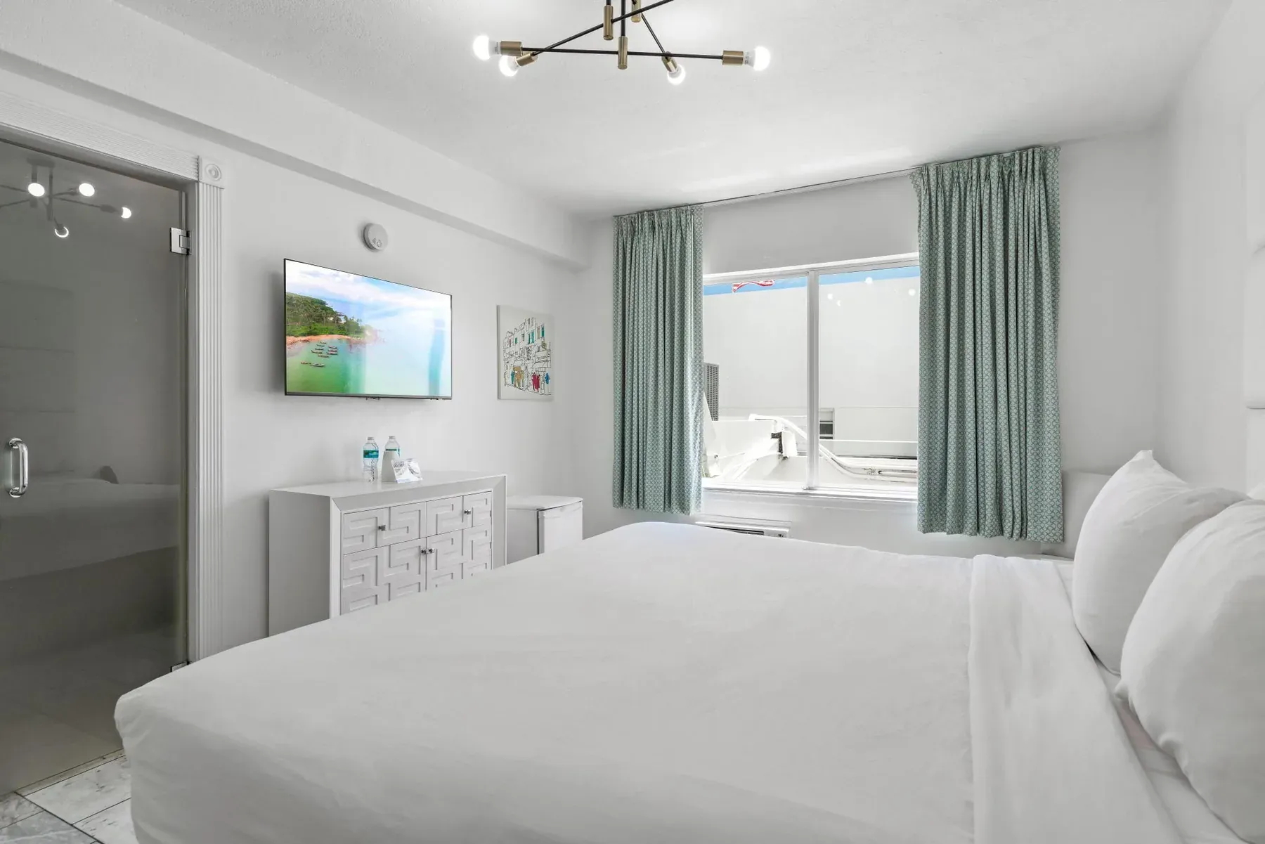 The room from the foot of the bed, television, dresser and chandelier